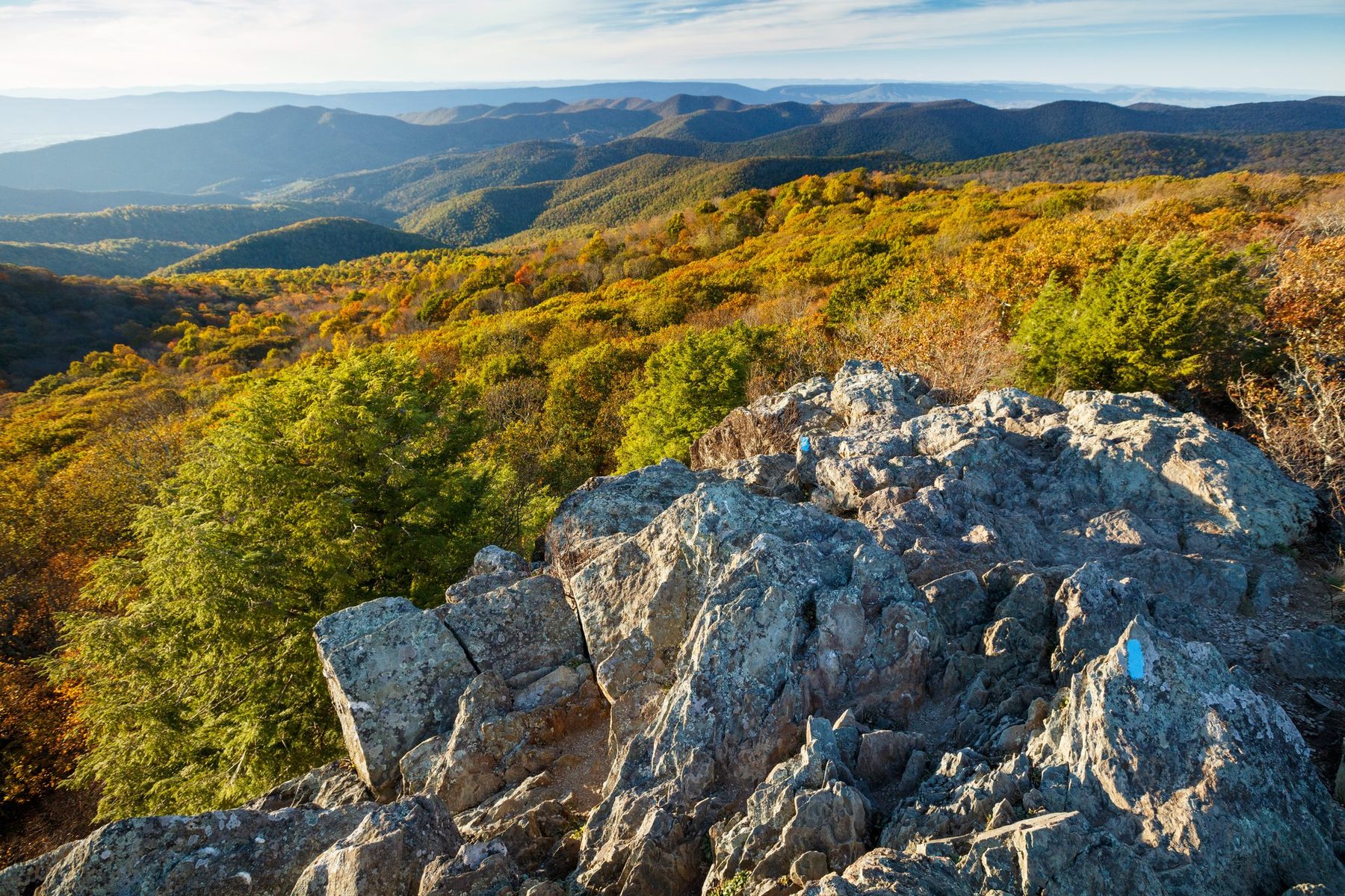 Late-afternoon light on a rocky Shenandoah summit, fall foliage and layered Blue Ridge ridgelines rolling to the horizon